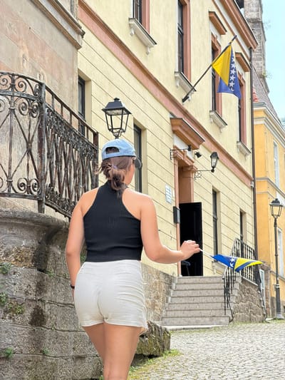The image depicts a young woman holding a small Bosnian flag on a street in Jajce, Bosnia and Herzegovina. She is wearing a black sleeveless top, white shorts, and a light blue baseball cap. Her hair is pulled back in a bun, with a purple clip visible. She is standing on a stone staircase, with a wrought-iron railing to her left. Behind her, a yellow building with brown trim and black windows rises up. The building has several floors, and a traditional street lamp is mounted on the wall. Another, larger Bosnian flag is mounted on the building, to the left of the woman. The flags are the national flag of Bosnia and Herzegovina, featuring a yellow triangle and white stars against a blue background. The setting appears to be an old European town, with cobblestone or stone pathways and historic architecture. The lighting suggests it is daytime, and the sky is visible at the top of the image, appearing overcast. The woman's pose and the flag suggest a moment of national pride or a visit to a historical site.