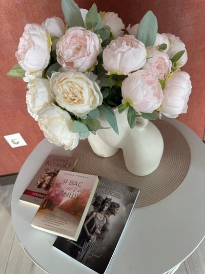 This media file captures a still life arrangement on a white table with a reddish-brown surface underneath. The foreground features a bouquet of artificial peonies, predominantly white and pale pink, with green foliage. To the right of the flowers, on the white table, are three books. The book closest to the viewer has a pale pink cover with an illustration of trees and has Russian text. The text on this book reads "ЕЛЕНА РОНИНА Я ВАС не_у слышу". A second book, partially visible behind the first, has a red spine and the Russian text "ПАУЛО КОЭЛЬЮ АЛХИМИ" visible on its cover. The third book, lying flat and angled, has a dark cover with an illustration of two figures and the Russian text "МНОГОЛИКАЯ ЛЮБОВЬ Анатолий Лагунов 18+". Beneath these books is a beige, woven placemat, and peeking out from behind the flowers and placemat is a portion of a white, stylized bust or figurine. The overall impression is one of a domestic or quiet setting, possibly a living room or study, given the presence of books and decorative items. The lighting suggests it might be daytime, possibly indoors. There are no discernible weather cues or explicit location cues beyond the Russian text on the books.