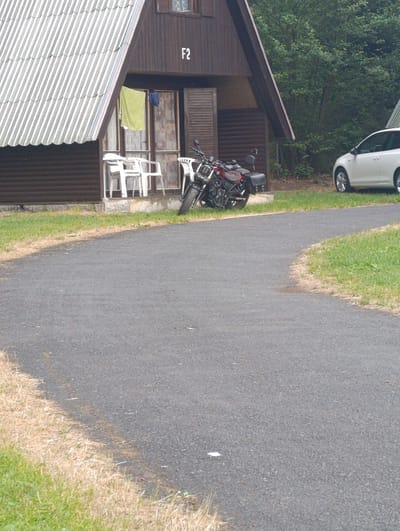 The image depicts a scene in a rural or semi-rural setting, likely near a small building or cabin complex. The building in the foreground, characterized by a steeply pitched corrugated roof and dark wooden siding, has the designation "F2" visible on its exterior. In front of the building, several white plastic chairs are arranged on a concrete base. A red and black motorcycle is parked to the right of the chairs. A light-colored car is visible to the right of the motorcycle, parked on what appears to be a paved surface that curves away into the foreground. The environment includes grass and some dry vegetation along the edge of the paved area, and dense foliage of trees in the background. There are no people visible in the image. The location is identified as Babylon, Czechia.