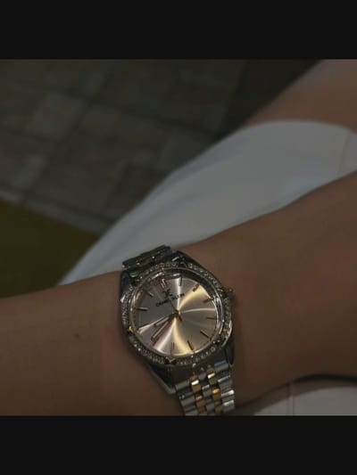 A close-up shot features a silver and gold Daniel Klein watch with a diamond-encrusted bezel, worn on a person's wrist. The watch face is silver with thin, rose-gold hour markers and hands. The background shows a person wearing a white garment and an out-of-focus tiled surface. The location in Bielawska Wieś, Poland, is not discernible from the image.