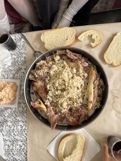 The image depicts an overhead view of a communal meal served on a table in a domestic setting within the city of Breznitsa, Bulgaria. Central to the scene is a large, round metal pan containing a main course of meat and rice. The meat, likely lamb, is presented as pulled or shredded pieces mixed with lighter-colored long-grain rice, with some areas showing a darker, possibly spiced, rice preparation. Visible within the dish are several bone fragments, including a prominent long bone on the right side.
Surrounding the pan are several slices of white bread; one slice in the upper center is whole, another to its right is partially consumed with a large bite taken from it, and a third, thinner slice is visible in the upper right. Another slice of bread, partially eaten, rests on a white paper napkin in the lower right.
To the upper left, a clear plastic cup filled with a dark, carbonated beverage and ice is placed on a white paper towel. A silver spoon lies adjacent to this cup and the main pan. Further left, a rectangular transparent plastic container holds several light-colored, irregularly shaped items, possibly cookies or biscuits.
In the upper background, partially visible legs and lower torsos of at least two individuals are present, suggesting multiple people are seated around the table. One person wears white leg coverings and shoes, while another wears dark pants. In the lower right foreground, a hand, likely a child's, holds a black cup containing a dark beverage.
The table surface is primarily covered with a light brown, possibly paper or disposable, tablecloth, exhibiting several dark, circular stains. A section of the table on the far left is covered by a white fabric tablecloth with a repeating black floral or leaf pattern.