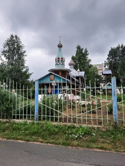 The image shows a wooden church with multiple domes and crosses, located in the city of Zelenogorsky, Russia. A green metal fence runs across the foreground, separating the viewer from the church and a garden with pink and white roses. Several trees are visible behind the church, and a multi-story building stands in the background. The sky is overcast and gray. On the left side of the fence, there are three lamps attached to a pole. Within the fenced area, near the church, a group of people are standing. One person appears to be wearing a green dress, another a yellow shirt, and a third is partially obscured. The church itself has blue trim and a wooden exterior.