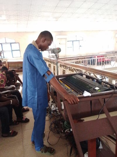The image shows a man, dressed in a blue uniform, adjusting a sound mixer at a location in Yenagoa, Nigeria. He is standing on a wooden platform, adjusting a large sound mixer with multiple knobs and sliders. He appears to be adjusting the levels on the mixer, with his hand placed on the side of the equipment. In the background, there are several other individuals sitting on a lower platform with a railing, and there is a large ceiling fan mounted to the left of the sound mixer. The scene is in a well-lit room with tiled flooring. There are numerous electrical cords and equipment on the floor around the sound mixer. The image captures a moment of technical activity, likely during a gathering or event.