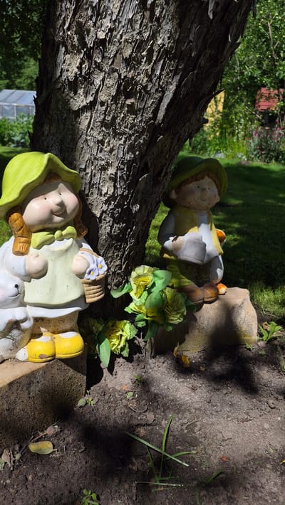 A frontal medium shot captures two garden gnome figurines positioned next to the trunk of a mature tree. The gnome on the left, dressed in a light green hat and apron over a white outfit, holds a basket with blue flowers. A white rabbit figurine is partially visible to its left. The gnome on the right wears a green hat and yellow vest over a white shirt, holding a grey bucket. Both gnomes have tan skin and dark hair. They are situated on dark soil, with a few artificial yellow flowers placed between them. The tree trunk is textured and dark. In the background, a well-maintained lawn is visible, along with some distant buildings and greenery. The scene is illuminated by sunlight, casting shadows on the ground.