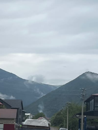 The image shows a cloudy sky over a mountainous landscape in the village of Krasna, Ukraine. In the foreground, there are several buildings and a street. On the left, a building with a dark grey roof and a brown-red lower section is visible. Adjacent to it is another dark grey building with multiple windows. In the center, a white object, possibly a vehicle or structure, is parked on a dark surface, and a small yellow figure is seen near it. Further to the right, a building with a dark pitched roof and glass windows is present. Utility poles with wires run through the scene. In the background, the lush green slopes of mountains are covered in parts by mist and clouds. Smoke plumes are visible rising from the mountainside, suggesting human activity such as heating or cooking. The overall atmosphere is overcast and slightly hazy.
