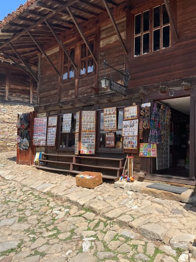 The image depicts the exterior of a souvenir shop in the historic town of Zheravna, Bulgaria. The shop is housed in a traditional wooden building with a tiled roof and exposed wooden beams. In front of the shop, various souvenirs are displayed on wooden boards and racks, including bags, jewelry, magnets, and postcards. A cobblestone street leads up to the shop entrance, which is open, revealing more merchandise inside. A cardboard box filled with colorful items is placed on the street in front of the shop. A simple wooden bench is situated in front of the display windows. The overall scene is representative of a charming, historical tourist attraction.