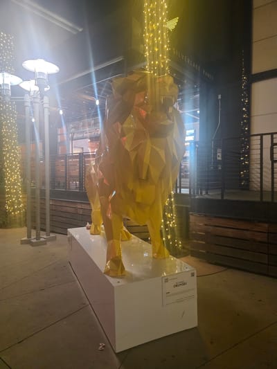 An outdoor night-time scene in Burbank, California, features a large, geometric, gold-colored lion sculpture standing on a white pedestal. The sculpture is illuminated by string lights that cascade from above, creating a bright, festive atmosphere. In the background, the facade of a building is visible, with signage that reads "SILVERLAKE RAMEN." The area around the sculpture appears to be a walkway or patio, with concrete pavement and wooden fencing. The lighting is somewhat harsh, with strong highlights and lens flares from the lights, obscuring some of the background details. A small plaque is visible on the pedestal of the sculpture, indicating it is titled "LION" by artist RICHARD ORLINSKI.