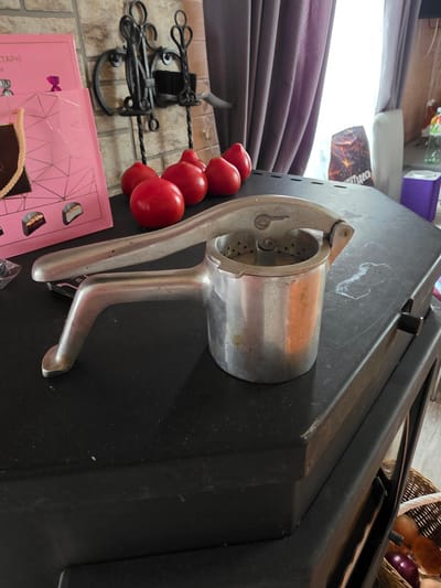 The image depicts an indoor scene, likely a home kitchen, located in Проводник, Russia. In the foreground, a metal potato ricer sits on a black surface. To the left of the ricer, a pink box with geometric patterns and some candies is visible, and a pink gift bag with a braided handle is also present. Behind the ricer, several red tomatoes are arranged. In the background, a brick wall is partially visible, with a metal fireplace tool set mounted on it. To the right, a purple object and a clear plastic bottle with a colorful label are on a surface. A window with purple curtains is also visible. The overall lighting suggests an indoor environment, possibly during the daytime.