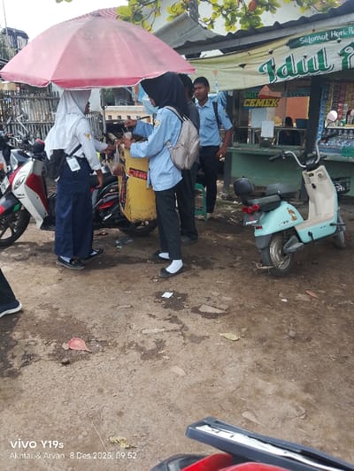 Three students in blue uniforms and white headscarves gather around a yellow cart beneath a pink umbrella. One student appears to be purchasing food from the cart. A male student in a similar uniform stands nearby. The scene takes place near a small shop with a banner reading "Selamat Hari Raya Idul..." and "MIE CEMEK". A light blue electric scooter is parked on the side. Several motorcycles are visible in the background. The ground is uneven and muddy. The photo was taken on December 8, 2025, at 09:52.
