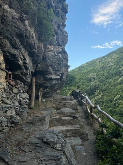 The image depicts a rugged, natural scene with a focus on rocky cliffs and lush green foliage. The perspective is from a low angle, looking upwards towards a steep, densely forested hillside that dominates the background. The sky above is a clear blue, with scattered white clouds, suggesting fair weather.
The foreground is characterized by a dramatic rock face, heavily textured with fractures and crevices. Large, weathered rocks and boulders are scattered across the lower part of the image, indicating a natural, untamed environment. A fallen log lies horizontally across a section of the cliff face, adding a sense of natural debris. Further down, there are visible remnants of what appear to be old industrial equipment, possibly abandoned mining or quarrying machinery, suggesting a historical human presence in this otherwise wild setting.
A rough, unpaved path runs along the top of the rocky embankment, with a primitive railing made of branches and logs. This railing appears to be an attempt to provide safety or guidance along the potentially hazardous terrain. The path leads towards the dense forest.
The overall impression is one of a remote and somewhat wild natural landscape, possibly a hiking trail or a scenic overlook. The lighting suggests it is daytime, likely late morning or afternoon, given the brightness and the shadows. The location cues, such as the type of rock and vegetation, combined with the provided location context of Golyamo Belovo, Bulgaria, point to a mountainous or hilly region. There is no visible text in the image.