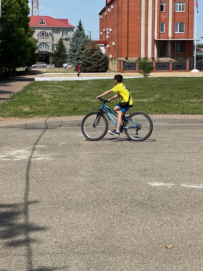 A young boy is riding a blue bicycle on an asphalt path. He is wearing a yellow t-shirt and blue shorts. The bicycle has black tires and a black frame. The boy is leaning forward as he rides, and his shadow is cast on the ground behind him. The scene appears to be a park or a recreational area, with green grass and trees visible in the background. There is also a building made of red brick in the background. The weather appears to be sunny, and the time of day is likely daytime. No emotions are discernible from the boy's posture. The visible text on the bicycle reads "FORWARD."