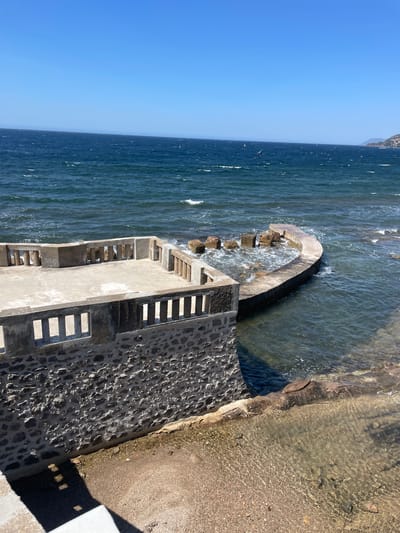 A paved viewing platform with a stone-clad base overlooks the Mediterranean Sea in Carqueiranne, France. The platform features a concrete floor and a low concrete balustrade with rectangular openings. Adjacent to the platform, a curved concrete seawall encloses a small section of the sea where rocks are visible. The water is a deep blue, with gentle waves and white foam. A sandy and pebbly shore is visible in the lower right of the image. The sky is clear and blue, with distant hazy landmasses on the horizon. There are no people or animals visible in the scene.
