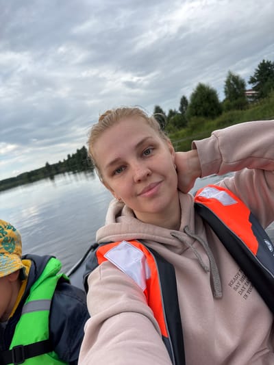 A woman with blonde hair pulled back in a bun and freckles across her nose and cheeks is taking a selfie from a boat on a cloudy day. She is wearing a light pink hooded sweatshirt and an orange life jacket over it. Her left arm is raised and resting behind her head, and she is smiling at the camera. To her left, a child wearing a yellow bucket hat with a green and blue pattern and a bright green life jacket is visible. The boat is on a body of water, likely a river or lake, with a tree-lined shore in the background. The sky is overcast with grey clouds. The location is the city of Anteplevo (Okhotinskoe S/P), Russia.