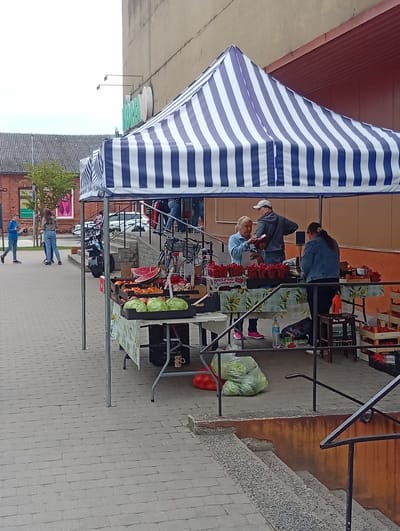 The image depicts an outdoor market stall selling fruits and vegetables, located on a paved walkway in front of a building. A striped blue and white awning covers the stall, which is laden with produce including strawberries, watermelons, and cabbages. Three individuals are present: two women and one man, seemingly engaged in transactions or discussions at the stall. In the background, other people are visible walking on the walkway, and a building with a brown facade and a grey roof is present. The environment appears to be an urban setting, possibly a small town or city square, given the paved walkway, buildings, and presence of parked bicycles and a scooter. The image was taken in Kuršėnai, Lithuania, on a cloudy day.