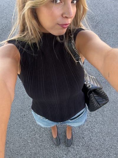 A female subject is depicted from a high-angle perspective, capturing a full-body selfie against a paved, dark gray ground surface. The individual has light-colored hair, dark eyes, and is looking directly at the camera. She wears a black, sleeveless, high-neck ribbed top, light blue denim shorts, and black, studded ballet flats. A black quilted shoulder bag with a silver chain strap is visible on her right side. A small, minimalist tattoo is present on her left forearm. The scene occurs outdoors in the city of Leiria, Portugal.