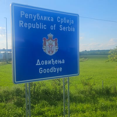 The image shows a blue border sign indicating the Republic of Serbia, with the Serbian coat of arms displayed in the center. The sign also reads "Довиђења" and "Goodbye" in Serbian and English. The sign is likely located at the border of Serbia in the Sid Municipality. The background features a green field with vegetation. The sky is light blue.