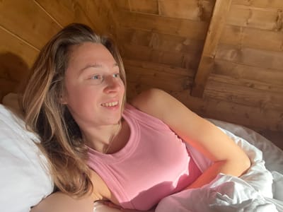In an interior setting within the city of Crkvine, Montenegro, a female individual is depicted lying in a bed. She is positioned on her back or side, with her head resting on a white pillow. Her long, light brown hair is loose around her head and shoulders. She wears a pink ribbed tank top and a thin silver-colored necklace. Her face is turned towards the viewer's right, with open eyes looking in that direction and a slight upward curve of the lips. Two faint, small, light-colored marks are visible above her left eyebrow. Her right arm is partially visible, resting on white bedding that covers the lower part of her body. The background consists of an inclined wooden ceiling or wall constructed from horizontal planks, with a prominent diagonal wooden beam. Strong natural light, consistent with direct sunlight, illuminates the scene from the right, casting distinct shadows.