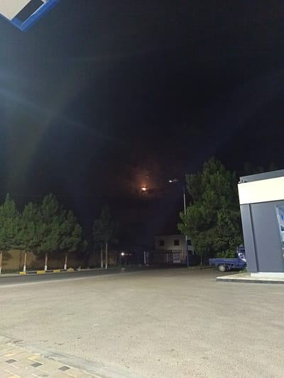 The image depicts an outdoor scene at night in the city of Guliston, Tajikistan. In the sky, a bright, reddish-orange circular object, likely the moon, is visible. A street light illuminates the area, casting light on a row of green trees lining a road. Further back, a light-colored building with dark windows can be seen. To the right of the frame, a modern building with gray and white sections stands, and a small blue truck is parked nearby. The foreground shows a paved area, possibly a road or parking lot, with a sidewalk made of patterned tiles on the left.
