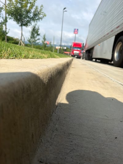This image shows a low-angle view of a concrete curb on a sidewalk. In the background, there is a red semi-truck and a white semi-truck, possibly stopped at a traffic light. The red truck has "6" visible on a sign behind it. There are trees and grass to the left of the curb, and the sky is cloudy. The scene appears to be along a road in a city, with the distinct signage of a truck stop or gas station visible in the distance. The overall lighting suggests it is daytime. While the image is taken in Chattanooga, United States, specific landmarks or street names are not identifiable from the visual information provided.