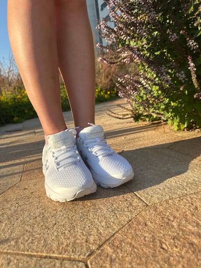 The image shows a person's legs and feet wearing white Under Armour Charged Gemini sneakers. The shoes are laced and have text on the tongue. The person is sitting or resting on a paved surface, possibly a sidewalk or walkway, made of large stone tiles. The background features a patch of green plants with small pinkish flowers and some yellow flowers. The lighting suggests it is daytime, with sunlight casting shadows on the ground. The scene appears to be outdoors in a bright, possibly warm environment. The location context is Givat Shmuel, Israel. The visible text on the shoes reads "CHARGED GEMINI".