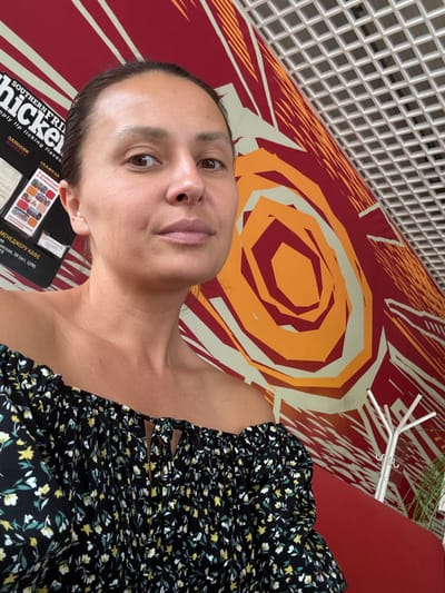A woman is depicted from the upper torso up, looking directly at the camera with a neutral expression. She has medium-toned skin, dark brown hair pulled back, and is wearing a dark, off-the-shoulder floral top with small yellow, white, and green flowers. Her right ear features a small pearl earring. The background to her left includes a black sign displaying partial English text "SOUTHERN FRI hicken" and "mply lip licking flavou," alongside Cyrillic text that translates to "Fill out the form" and "Cafe Manager" with an address reference "(stop. TSUM)". The wall behind her features a large, abstract graphic design in red, orange, and light beige geometric shapes and radiating lines. A white grid ceiling is visible in the upper right. A red bench or couch, a white minimalist coat rack, and some green foliage are partially visible in the lower right background. The scene is located in Krilovo, Russia.
