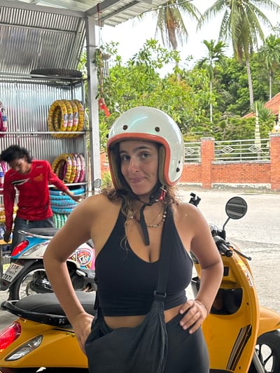 The image depicts a daytime scene at a scooter rental or repair shop in Baan Nai Wok, Thailand. In the foreground, a woman with medium-length, curly brown hair wears a white retro-style motorcycle helmet with an orange trim and a black chin strap. She is dressed in a black halter-neck crop top and black pants, with a black cross-body bag slung over her shoulder. Multiple layered necklaces and a bracelet are visible. She stands with her hands on her hips, looking directly at the camera with a slight smile. A yellow scooter is positioned to her right, and part of a white and blue scooter is visible behind her to the left. In the background, to the left of the woman, a man with dark hair, wearing a long-sleeved red shirt and blue jeans, is partially obscured as he appears to be working near stacks of new motorcycle tires. These tires are displayed on corrugated metal shelves under a corrugated metal roof structure. Beyond the shop area, lush tropical foliage, including palm trees, is visible, bordered by a low brick wall with a diamond pattern. The ground beneath the subjects appears to be a paved surface.