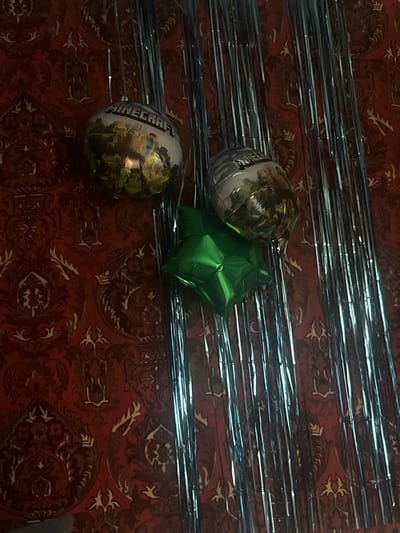 The image depicts a close-up, angled view of a wall decorated with a red, patterned tapestry. Hanging against the tapestry are several balloons and a curtain of silver tinsel strips. One balloon is round and features an image of characters from the video game Minecraft, along with the game's logo. Another balloon, also round, is metallic gold with some indistinct imagery. A green, star-shaped balloon is positioned below the other two. The tinsel strips hang vertically, creating a shimmering backdrop. There are no people visible in the image. The location is specified as Қанабек, Kazakhstan.