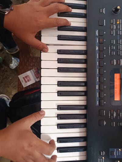 An overhead view depicts a person's hands actively playing an electronic keyboard in Kawungluwuk, Indonesia. The keyboard features white and black keys, with a dark gray control panel on the right containing an orange-backlit LCD screen displaying "F#th", "P=2", "SCL", "O", and "and". Numerous buttons are visible, including labels such as "RHYTHM," "GUITAR," "ORCH," "BRASS," "SYNTH," and "TEMPO." The upper hand, positioned over the higher octave keys, wears a dark smartwatch on the wrist. The lower hand is positioned over the lower octave keys. To the left of the keyboard, on a dark brown woven surface, a red and white pack of Gudang Garam International cigarettes is partially visible, alongside a dark bag and out-of-focus footwear in the background.
