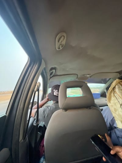 The interior of a car is visible, focusing on the back seat. Two passengers are present. One wears a striped shirt and has a beige cloth draped over them, while the other wears a gray shirt and a black cap with a white design. A cell phone is held in the upper right corner. The car's roof is gray, and the window shows an unclear view of land or sky. The headrest of the front seat is prominently displayed, obscuring some of the view.