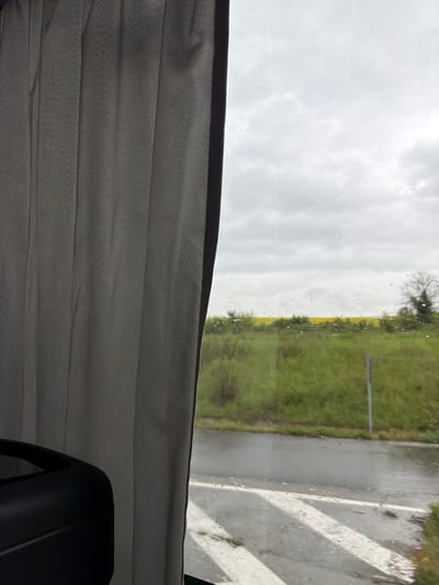 The image captures a perspective from inside a vehicle, looking out a side window during an overcast and rainy day. The view is presented as if the camera was held sideways, showing the scene rotated 90 degrees clockwise.
In the lower half of the frame, a pleated grey fabric, resembling a curtain or blind, is visible, indicating the vehicle's interior. A dark, possibly black, object, potentially part of a car seat or interior fixture, is partially visible in the very bottom right.
Through the window, numerous raindrops cling to the glass, suggesting ongoing or recent rainfall. Beyond the wet pane, the sky is uniformly grey and cloudy. The landscape consists of expansive green fields or grasslands, with subtle patches of yellow, possibly from blooming crops like rapeseed. Scattered trees, some appearing bare or with minimal foliage, punctuate the horizon. To the right of the view through the window, a wet, grey road or paved area is visible, marked with distinct white lines, which could be lane markings or a pedestrian crossing. The scene evokes a sense of travel or observation during rainy weather in a rural or semi-rural setting. The location context provided is Rogozinovo, Bulgaria. No text is visible in the image.