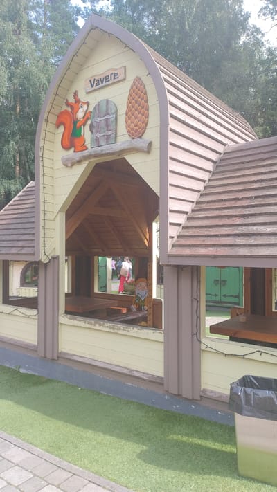 The image depicts a whimsical, light-yellow wooden structure resembling a playhouse or a small shelter, set against a backdrop of trees. The building has a gabled roof with brown, plank-like shingles and decorative trim. The front of the structure features an open archway, revealing an interior with wooden beams and tables.
Dominating the upper facade is a painted sign reading "Vavere" above a depiction of a squirrel holding a nut, next to a stylized pinecone and a small, rustic door. To the right of the entrance, a small gnome figurine is visible, perched on a shelf with a measuring scale behind it. In the background, through the open structure, a person in red pants and a white shirt can be seen, along with a pair of green double doors.
The ground in front of the structure is covered with artificial turf, bordering a paved walkway. To the far right, a trash can with a black liner is situated on the turf. The overall setting suggests a park, playground, or recreational area, likely in Kroņauce, Latvia, given the context. The scene is well-lit by daylight, and the atmosphere appears cheerful and family-friendly.