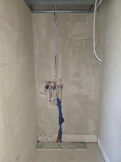 The image shows an unfinished interior wall in Łagiewniki Nowe, Poland. A metal ceiling grid is visible at the top, with white electrical conduit running vertically down the wall. In the center of the wall, a plumbing manifold with multiple pipes and valves is exposed, with red and blue pipes emerging from the bottom. The wall itself is plastered and appears to be in a state of construction or renovation, with visible patches and an uneven texture. The floor is concrete, with some tile debris and two white tiles laid flat. The overall scene depicts a utility area within a building under construction.
