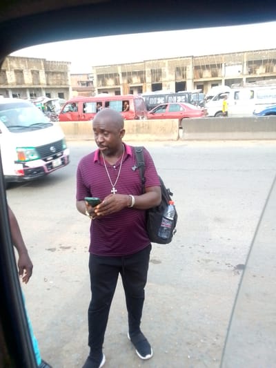 A man is standing on a street in Ihiala, Nigeria. He is holding a smartphone and is wearing a purple striped shirt, black pants, and black shoes. He has a backpack and a cross necklace. A white van and other vehicles are visible on the street behind him. Buildings are visible in the background.