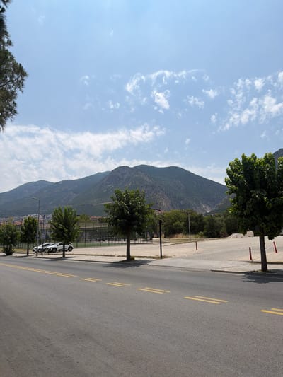 A landscape view shows a paved road in the foreground with yellow double lines marking lanes. To the left, a few trees line the road. Beyond the road, a fenced sports court with green turf is visible. In the background, a mountain range dominates the horizon under a blue sky with scattered white clouds. Several white cars are parked on the side of the road. The scene depicts an outdoor environment with elements of urban infrastructure and natural landscape. The overall impression is one of a sunny day in a town located near a mountainous region.