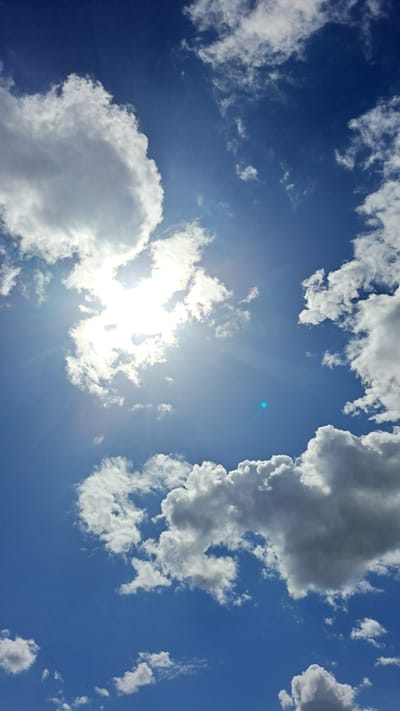 A wide-angle, low-angle shot of a blue sky with scattered clouds. The sun is visible, partially obscured by clouds, casting a bright glare and visible sun rays across the upper left quadrant of the image. Several large, puffy white clouds are present, with some darker grey clouds towards the bottom right. A small, faint blue lens flare is visible in the center right of the frame. The image appears to be a photograph taken outdoors. The scene does not contain any people or specific objects that would indicate a location within Marcinowice, Poland.