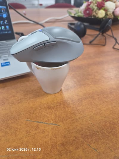 The image, taken in Gara Elin Pelin, Bulgaria, captures an indoor workspace scene on June 2, 2026, at 12:10 PM. The primary focus is an ergonomic computer mouse, light gray and white with a blue indicator light, positioned on a white ceramic mug. The mug itself shows prominent brown stains, suggesting it has been used for coffee or tea.
Partially visible beneath the mouse and mug is a white laptop, identifiable by a visible "Intel Core" sticker on its palm rest. The laptop screen displays various application icons. To the upper left, a vibrant bouquet of artificial or dried flowers, featuring shades of pink, red, cream, and green, sits in a dark container. Behind the flowers, a black, minimalist stand or holder is present. A dark cable runs across the table surface.
The items are arranged on a textured, mottled brown-orange surface, which appears to be a worn wooden or laminate tabletop with several prominent scratches and scuff marks, indicating frequent use. No people are visible, but the arrangement suggests a personal workspace. The timestamp "02 июня 2026 г. 12:10" is visible in the bottom right corner of the image.