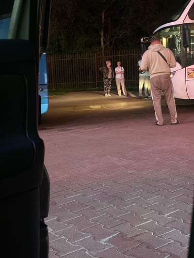 The image depicts an outdoor scene at night, in what appears to be a public area, possibly a bus stop or parking lot, in Terespol, Poland. In the foreground, a dark, reflective object, likely the interior of a vehicle, partially obscures the view on the left. The ground is paved with interlocking red bricks, suggesting an urban environment.
In the middle ground, several individuals are visible. On the right, a person wearing a light-colored hoodie and grey sweatpants stands facing a white bus. This person has a dark bag slung across their chest. To the left of the bus, three other individuals are gathered. Two women, one in a patterned jacket and dark pants, and another in a white t-shirt and light-colored trousers, are facing away from the bus and towards the viewer. A third person, partially obscured by the two women, is also present.
The background shows a dark fence and trees, indicating a more natural or green space adjacent to the paved area. The lighting suggests artificial illumination from streetlights or the bus itself, creating shadows and illuminating the scene in a low-light environment. There are no overt signs of distress or conflict, and the individuals appear to be waiting or interacting casually. The overall impression is one of a quiet, nocturnal scene in Terespol.