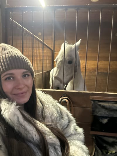 The scene takes place within an indoor stable or equestrian facility in Kalinovo, Russia. In the foreground, a female subject with long dark hair, wearing a light brown knitted beanie and a light-colored faux fur coat, takes a selfie. Her face is partially visible, displaying a slight smile. Behind a set of vertical metal bars, a white or light grey horse is visible. The horse's head and neck are positioned above a wooden stall partition, and it wears a dark bridle or halter. The horse appears to be looking directly towards the camera. The background consists of wooden walls, indicative of a barn or stable interior. An overhead light source is prominent at the top center of the image. To the right, below the horse's head, a brown wooden structure and some dark fabric pockets or bags are visible.