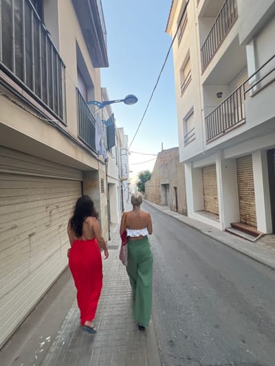 Two women are walking down a narrow street in L'Escala, Spain. The woman on the left is wearing a long, flowing red dress and blue sandals. The woman on the right is wearing a white crop top and green wide-leg pants. They are walking away from the camera, down a street lined with buildings. The buildings have balconies and shutters. There is a street lamp visible on the left side of the street. The street is paved with cobblestones. The sky is clear and blue.