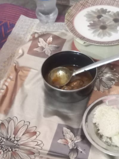 The image shows a meal setup on a floral tablecloth. A plate with a daisy pattern sits next to a black pot containing a curry, with a metal spoon resting inside. Adjacent to the pot is a clear bowl filled with white rice. A water bottle is also visible. The location is specified as Orai, India.