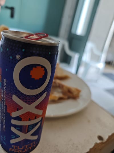 The image primarily features a vibrant can of beverage, positioned prominently in the foreground and slightly angled. Condensation droplets are visible on its surface, indicating it is cold. The can's design transitions from deep blue and purple at the top to reddish-orange at the bottom, overlaid with a pixelated pattern in various shades of blue, red, and turquoise. The brand name "OKIXO" is displayed in large white letters, with a pixelated orange square filling the center of the first 'O'. Smaller white text along the side reads "100% NATURALLY FREE" and "WITHOUT PRESERVATIVES". Towards the bottom, the flavor "TUTTI FRUITY" is visible. A red pull tab rests on the can's silver top.

In the blurred mid-ground, a white plate holds several pieces of golden-brown food, resembling pastries, crepes, or a type of baked or fried good. The can and plate rest on a light-colored surface, possibly a table or countertop, which shows some texture and dark markings in the bottom right corner. The background is a soft blur of an indoor setting, revealing light-colored objects that could be furniture or window frames, and a muted turquoise wall or partition. The bright lighting suggests a daytime scene. No individuals are visible, and the overall impression is a casual moment with a beverage and snack.