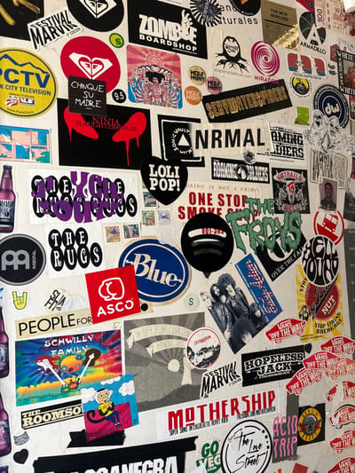 The image is a collection of stickers covering a surface. There is a diverse assortment of sticker designs ranging from logos, band names, slogans, and artwork. Some notable brands include Vans, Spotify, Blue, Meinl, and ASCO. Other stickers are artistic, featuring characters, text-based designs, and references to skateboarding. Some stickers seem vintage, while others are more contemporary.