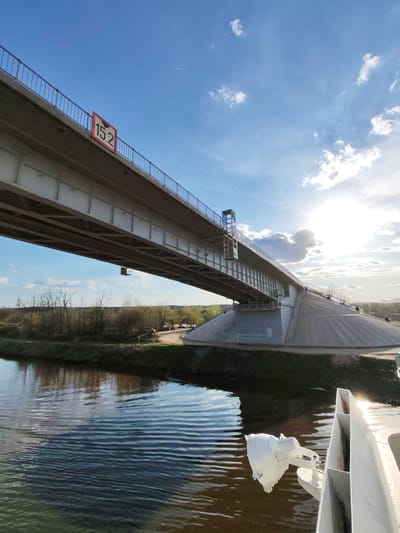 The image depicts a low-angle view of a bridge over a body of water. A sign with "15.2" is visible on the bridge. Trees and some grass are visible in the background, along with a road that looks like it is being used to park some vehicles. The weather appears to be sunny with a few clouds. Some of the ship's hardware is visible in the foreground. The location is Podosinki, Russia.