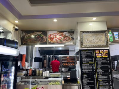 The image depicts the interior of a commercial food establishment, identified as a döner kebab and pizza shop in Tuttlingen, Germany. The scene focuses on the serving and kitchen area.

In the center-left, a male employee, wearing a red shirt and a dark baseball cap, stands with his back to the viewer, operating kitchen equipment. Visible behind him are a vertical rotisserie with a döner kebab spit and a horizontal rotisserie displaying multiple roasted chickens. Above this area, a large stainless steel exhaust hood is installed. The counter in the foreground features various food preparation items, including containers with fresh salad and vegetables, bottles of sauces (including soy sauce and sriracha), and a cash register system. To the far left, a tall black commercial refrigerator with a clear glass door showcases beverages like Red Bull and bottled water.

The upper wall features two large framed images: one displaying a large döner sandwich/wrap and another showing a pizza. To the right, a gold-framed artwork containing three circular elements with Arabic calligraphy and a clock face is mounted on a white tiled pillar. Below this artwork and adjacent to the kitchen area, a prominent black menu board with white text lists various German and Turkish dishes, including "Döner Teller," "Pizza," "Burger," and "Pide," along with their prices. An advertisement for Sprite and Ayran is positioned above the menu board. A white dome security camera is mounted in the upper left corner of the ceiling. The ceiling is white with recessed lighting and features a light purple accent border. The walls are partially tiled in white and black.