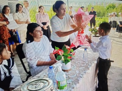 In Karagana, Uzbekistan, a group of people are gathered outdoors. In the foreground, a young boy in a light blue shirt and dark pants stands facing away from the camera. He is interacting with an adult woman who is holding a large, bright pink floral arrangement. The woman is wearing a white t-shirt and appears to be handing the arrangement to the boy.
To the left of the boy, a young girl wearing a black vest over a white shirt with a distinctive emblem is seated, looking to her right. Beside her, a woman in a white blouse is also seated and looking to her right, her arms crossed.
In the background, several other individuals are visible. Some appear to be adults, while others are likely students, possibly girls in school uniforms with white shirts and dark skirts. They are standing in rows, facing towards the main activity. The environment suggests an outdoor event, possibly a school gathering or celebration, with a backdrop of greenery and possibly some simple fencing or banners. A table covered with a patterned cloth is set up, displaying a vase of red and green flowers, a bottle of water, and a spray bottle. The ground is paved with what appears to be cobblestones or paving slabs.