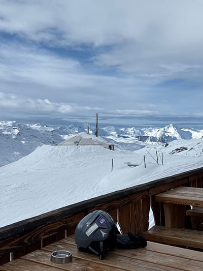 The scene presents a snow-covered mountain landscape in Saint-Martin-de-Belleville, France, viewed from an outdoor wooden terrace. In the foreground, a wooden table displays a silver-colored metal ashtray, a gray Atomic-branded ski helmet adorned with multiple stickers, and a pair of black gloves. A wooden railing runs across the lower portion of the frame, with a wooden bench partially visible to the right. Beyond the terrace, a vast expanse of snow-covered terrain features a prominent, dome-shaped structure, possibly a yurt, with a central vertical pipe and faint "COLUMBIA" branding on its side. Several dark, slender poles are embedded in the snow near this structure. The background consists of numerous snow-capped mountain peaks extending to the horizon under a sky with both blue patches and scattered white clouds. No individuals are depicted.