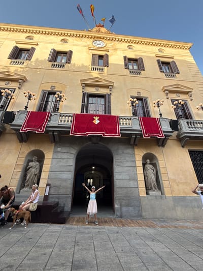 The image shows a wide, low-angle view of the exterior of a light yellow building, likely a public building in Mataró, Spain, given the crest displayed and flags. The building has a classical architectural style with multiple stories, visible windows with dark brown shutters, and balconies adorned with red velvet drapes bearing a coat of arms. Several ornate lamps with multiple bulbs are mounted on the balconies. At the top of the building, there's a decorative cornice, a clock face, and flags on poles.

In the foreground, a young person in a light-colored dress and a white cap stands with arms outstretched, facing the building, appearing to pose for a photograph. To the left, a few people are seated on a raised platform or steps. One woman wears a dark tank top and shorts, another woman wears a white tank top and a beige skirt, and a brown dog is near them. The ground in the foreground is paved with large, grey stone slabs. An arched entryway is visible in the center of the building, leading into a darker interior. The overall scene is set outdoors under a clear blue sky.