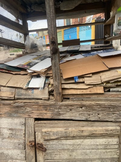 This image depicts a makeshift storage unit or stall located in Ojo, Nigeria. The lower half of the structure is constructed from weathered, horizontally arranged wooden planks, exhibiting rough textures and small boreholes. A closed wooden door with a rusty metal latch is situated in the bottom left. Above this wooden base, numerous flattened cardboard boxes and sheets are densely and irregularly stacked. Branding, including the letters "KO," is visible on some cardboard pieces. A central vertical wooden post, also weathered, features a rusty metal hook or latch mechanism. The upper sections of the structure incorporate horizontal wooden beams and sections of wire mesh. A transparent plastic bag containing unidentifiable items rests on the cardboard stack on the left. A small blue plastic object is embedded within the cardboard on the right. In the upper right corner, a translucent plastic container with a green lid sits on a narrow wooden shelf. The background, viewed through the mesh and gaps in the structure, reveals multiple buildings or walls painted in distinct blue, yellow, and brown colors, beneath an overcast sky. No individuals are present in the scene.