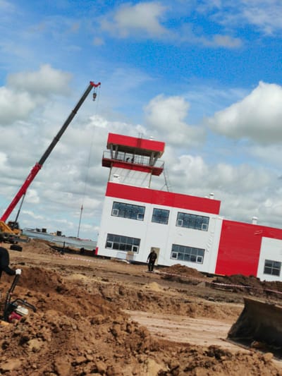 This is a medium shot, full-frame photograph taken outdoors on a cloudy day, depicting a construction site in the city of Arkalyk, Kazakhstan. In the background, a multi-story building with red and white sections and numerous windows is under construction. To the left of the building, a tall red and grey crane is positioned, with its boom extending upwards. In the foreground, a person wearing dark clothing and white gloves is operating a red and black construction machine, likely a compactor or vibratory roller, on a dirt mound. Another person in dark clothing stands near the entrance of the red and white building. The ground is uneven and covered with dirt and debris, indicating ongoing construction activity.