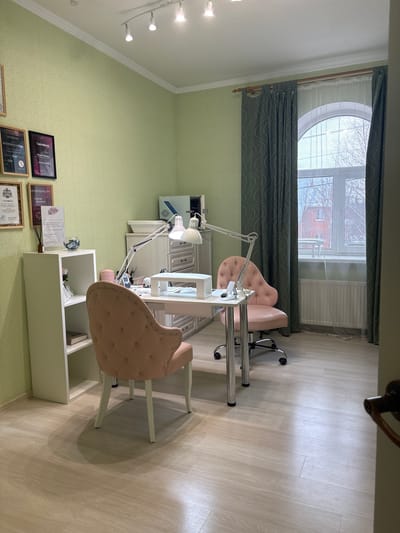 The image depicts an unoccupied interior room, functioning as a nail salon or beauty treatment area, located in Shul'gino, Russia. The walls are covered in light green, subtly patterned wallpaper. The flooring consists of light-colored wood-effect laminate panels. The white ceiling features a three-spotlight track lighting fixture.

On the right side of the room, a large, arched window is fitted with dark green, wave-patterned curtains and a sheer white curtain. Outside the window, bare tree branches and a distant red building are visible, suggesting an overcast or winter day. A white radiator is installed beneath the window.

Against the left wall, a white open-shelf unit displays a pink artificial flower, a reed diffuser, a glass bowl containing small items, and a book on the lower shelf. Above and to the left of this unit, multiple framed certificates or diplomas are affixed to the wall.

In the center of the room, a white manicure table with integrated drawers serves as the primary workspace. A white, dual-headed adjustable desk lamp is clamped to the table. On the table, a white nail dust collector with a hand rest, a smartphone in a stand, and various manicure tools and products are arranged. Two pink, tufted armchairs are positioned at the table: one stationary chair facing the table from the front, and one rolling, five-wheeled office chair on the opposite side.

Behind the manicure table, against the back wall, a light-colored storage cabinet features a grey countertop. On this countertop, a light blue and white sterilization unit and a white tray are visible. No individuals are present in the scene, and no actions or interactions are depicted.